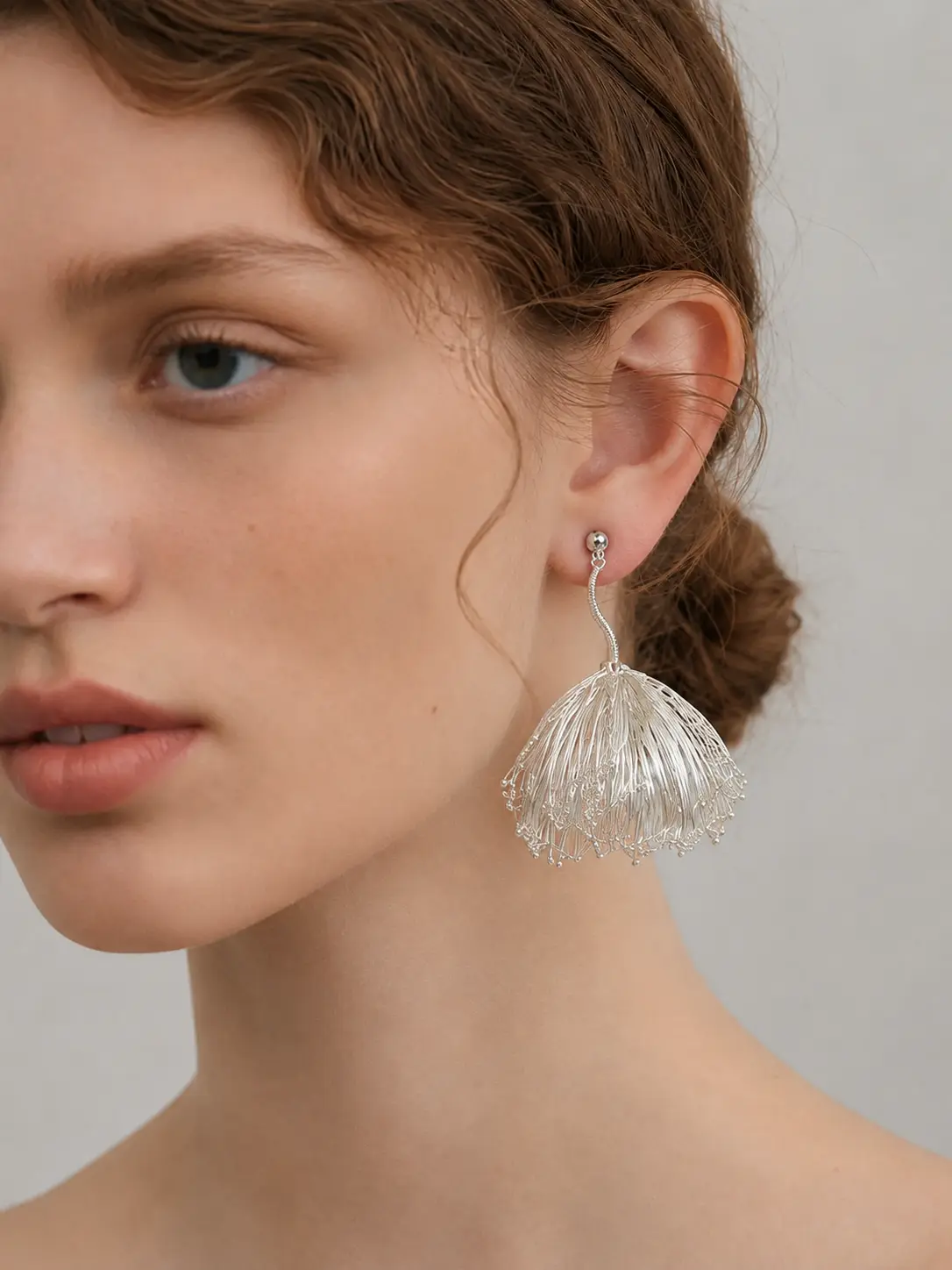 Close worn view of a silver-tone handwoven wire earring with a curved stem and sculptural bell-shaped drop